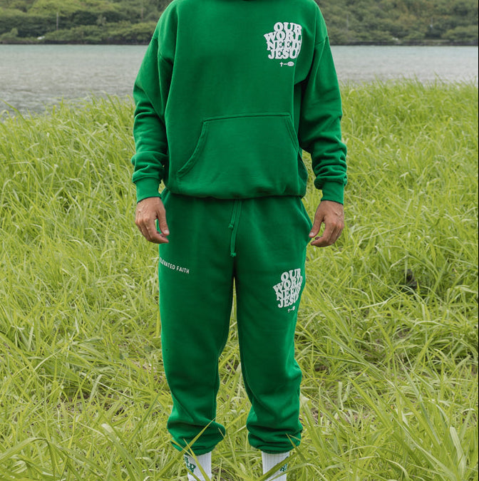 Man wearing a green hoodie and sweatpants standing in a grassy field with mountains in the background