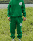Man wearing a green hoodie and sweatpants standing in a grassy field with mountains in the background