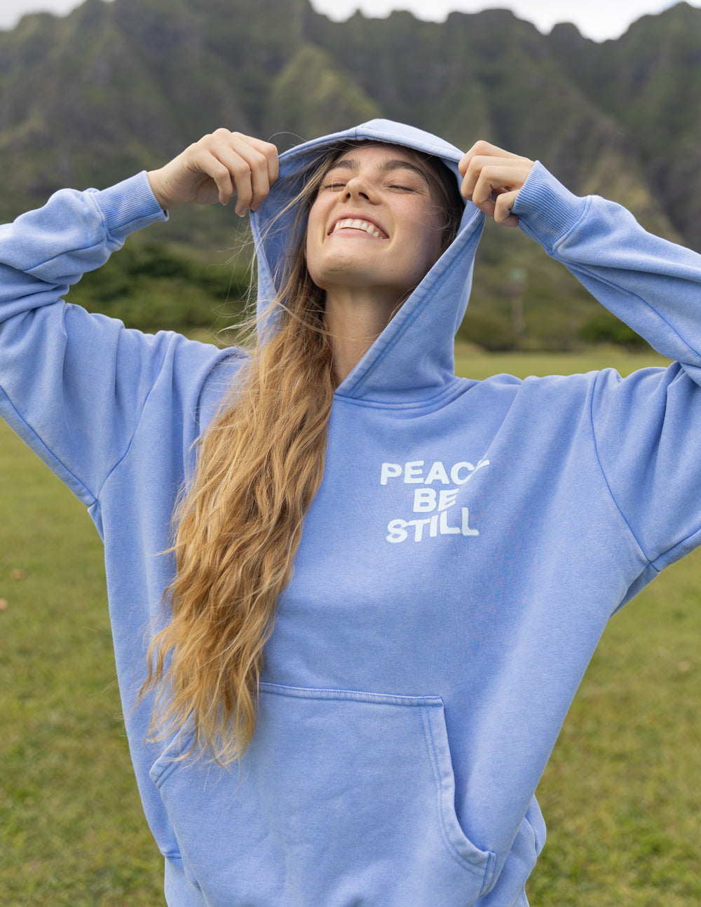 Model holding the hood of the blue Peace Be Still Hoodie outdoors.