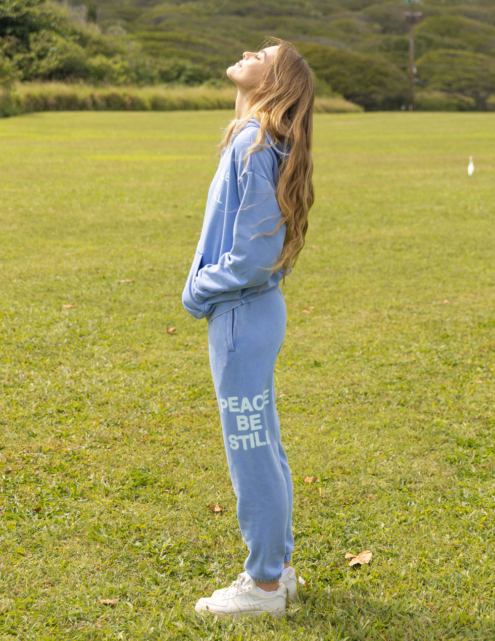 Side view of a model wearing blue Peace Be Still sweatpants with a coordinating sweatshirt outdoors.