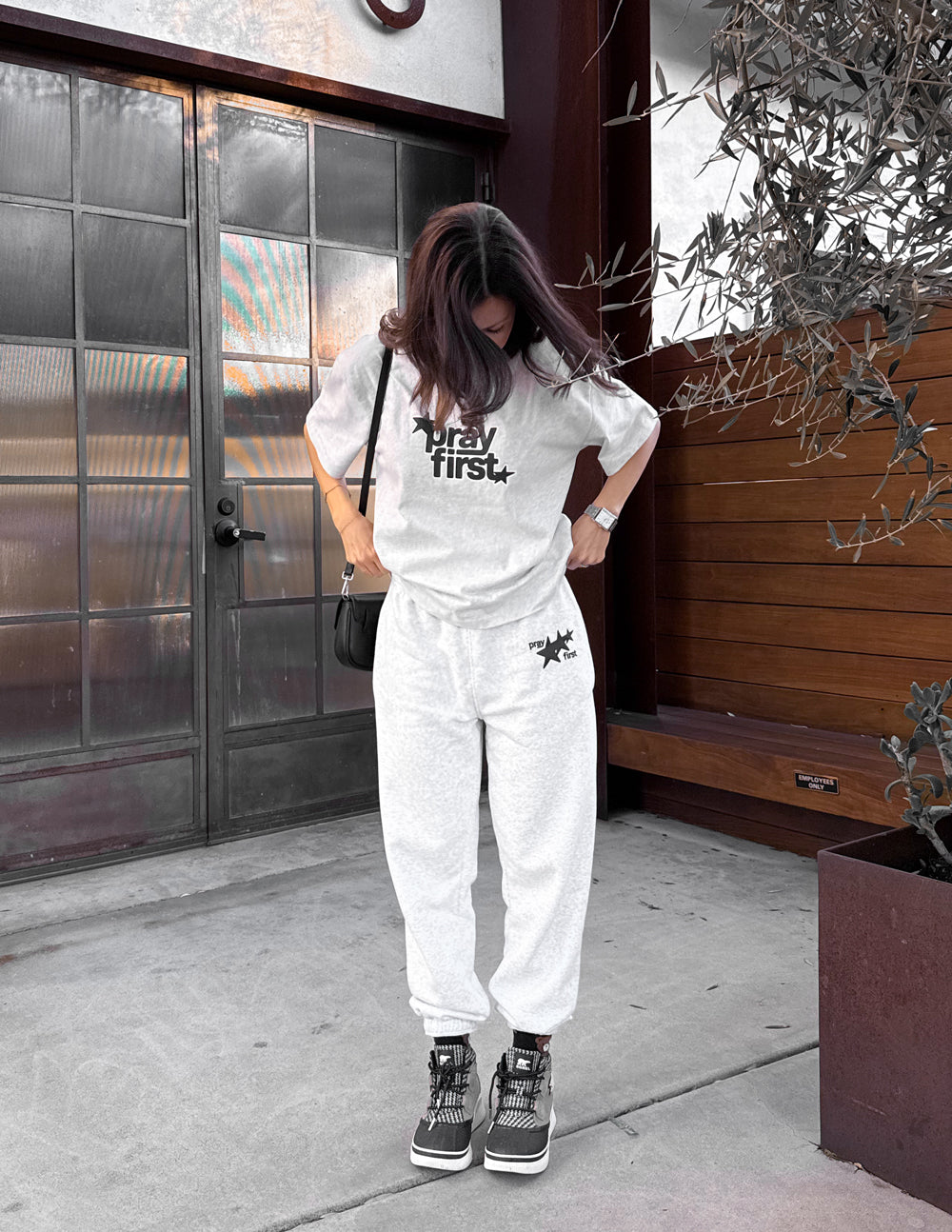 Model wearing the Pray First heather grey tee with light-colored pants and sneakers near a doorway