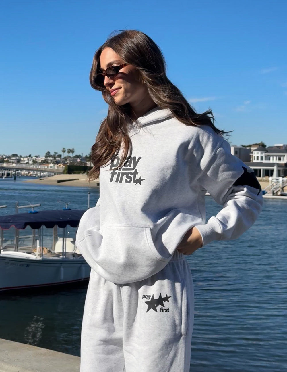 Woman wearing Pray First Heather Grey Hoodie with coordinating grey sweatpants beside the water
