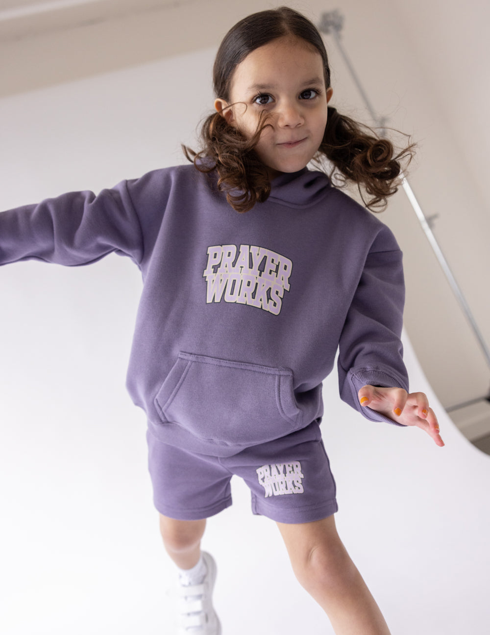 Child wearing the Eggplant purple Prayer Works hoodie with cream lettering and coordinating shorts.