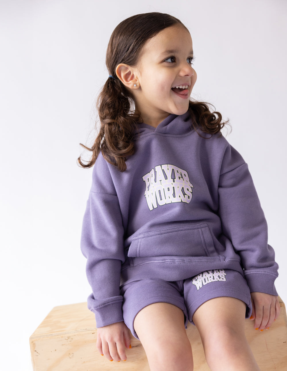 Child seated on a wooden bench wearing the purple Prayer Works hoodie and coordinating shorts.