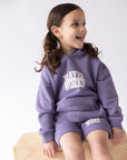 Child seated on a wooden bench wearing the purple Prayer Works hoodie and coordinating shorts.