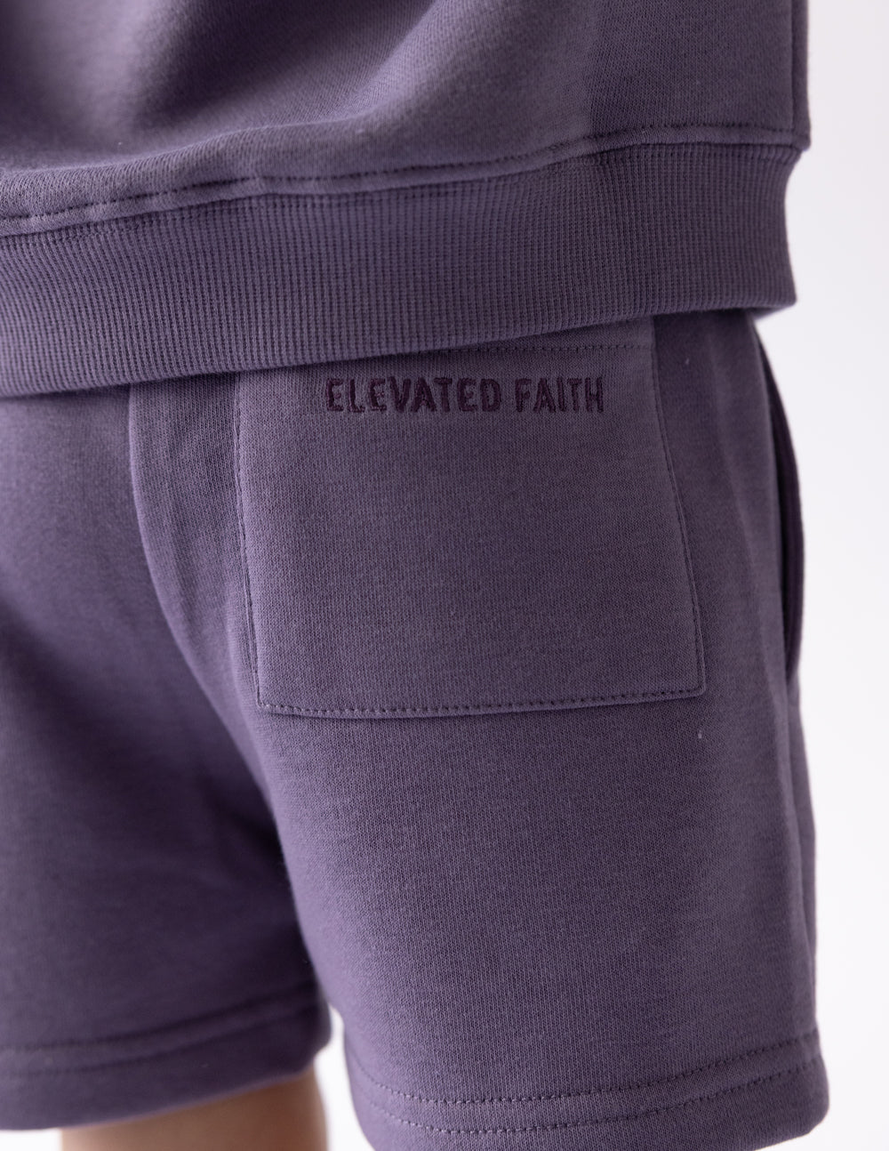 Close-up of the back patch pocket and Elevated Faith branding on eggplant kids shorts
