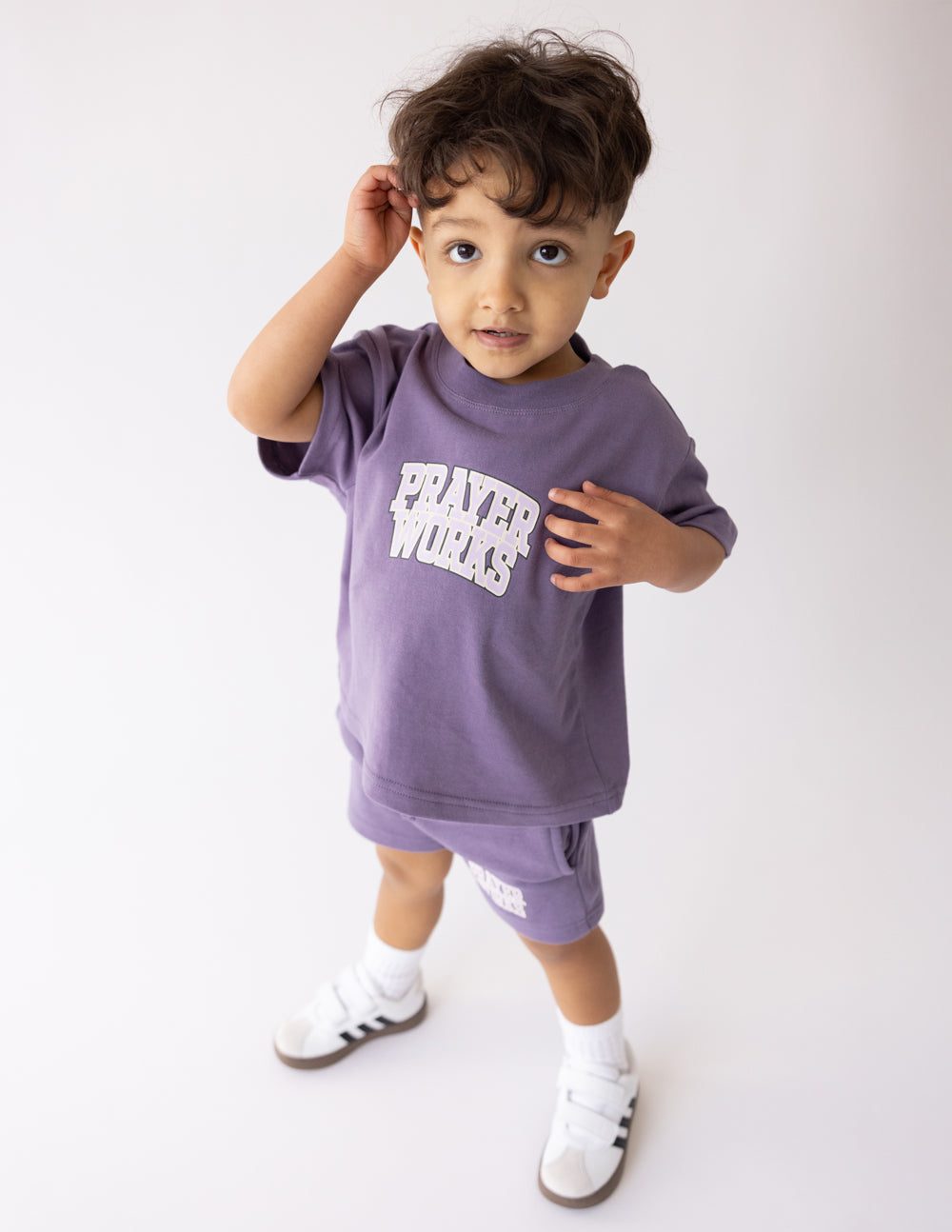 Child wearing the Prayer Works Eggplant Kids Tee with matching purple shorts and white sneakers