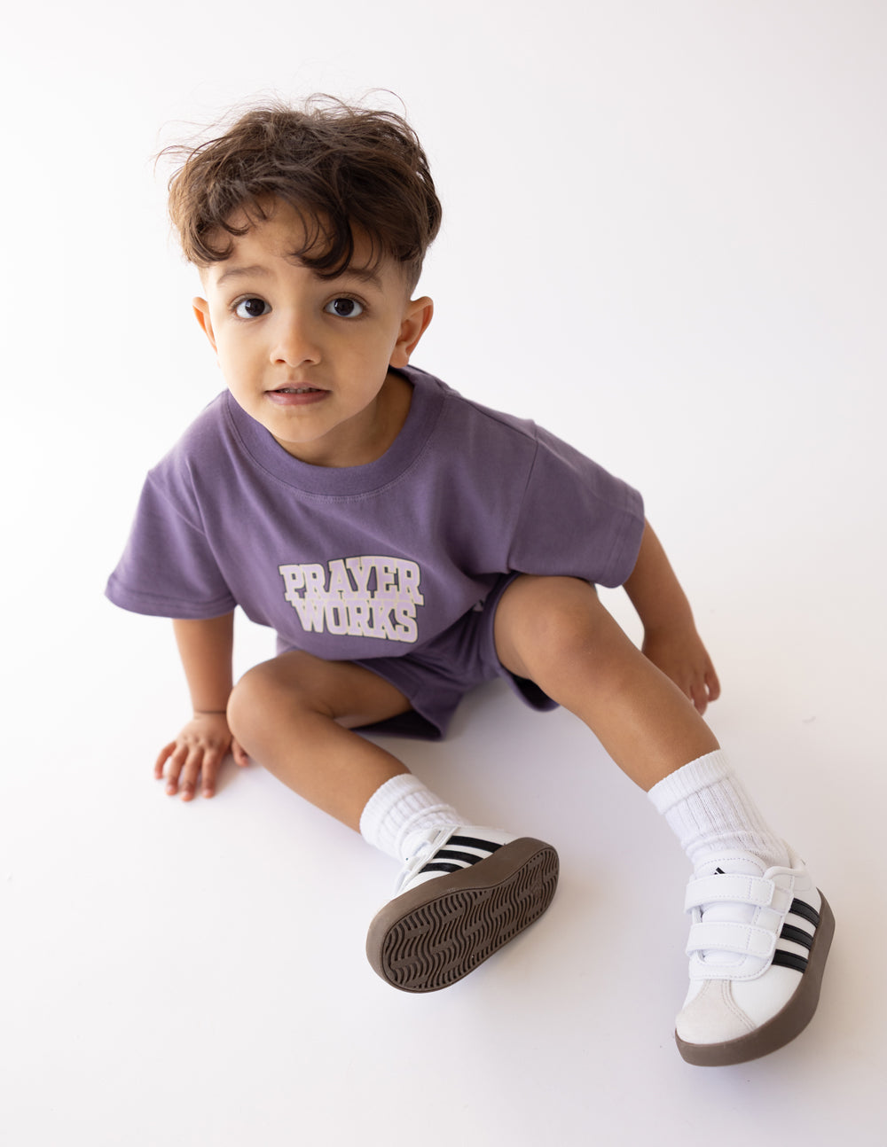 Child seated wearing the oversized Prayer Works Eggplant Kids Tee