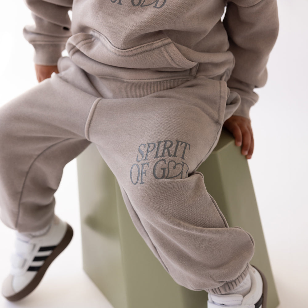 Child wearing a beige tracksuit with 'Spirit of G' text, sitting on a green stool.