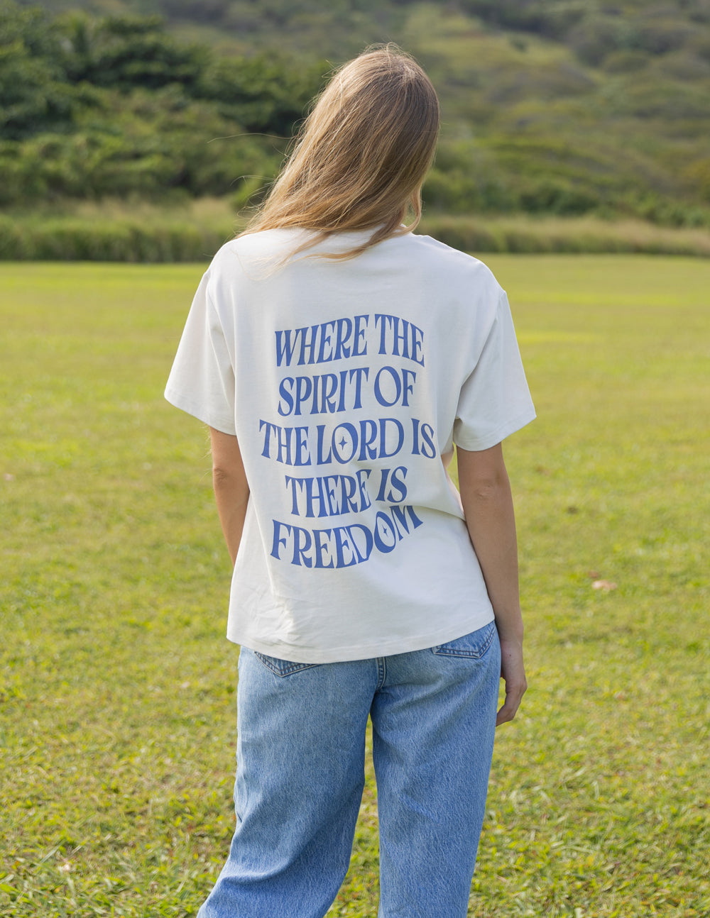 Back view of a model wearing the oversized Spirit of the Lord T-shirt with blue jeans in a grassy field.