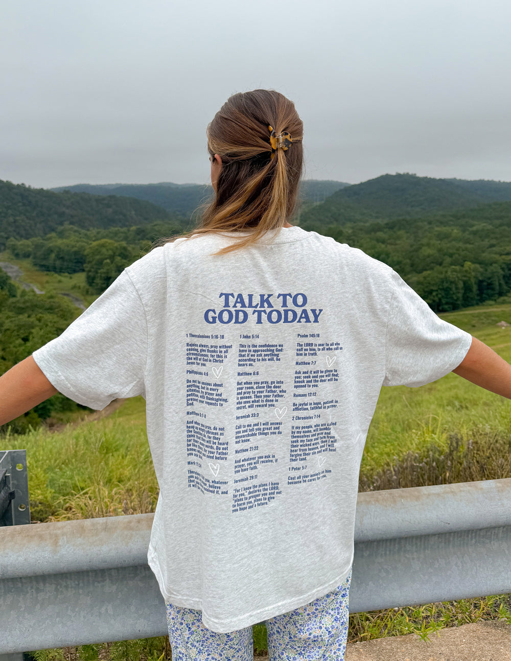 Back view of a woman wearing the heather grey Talk to God Today T-shirt overlooking green hills