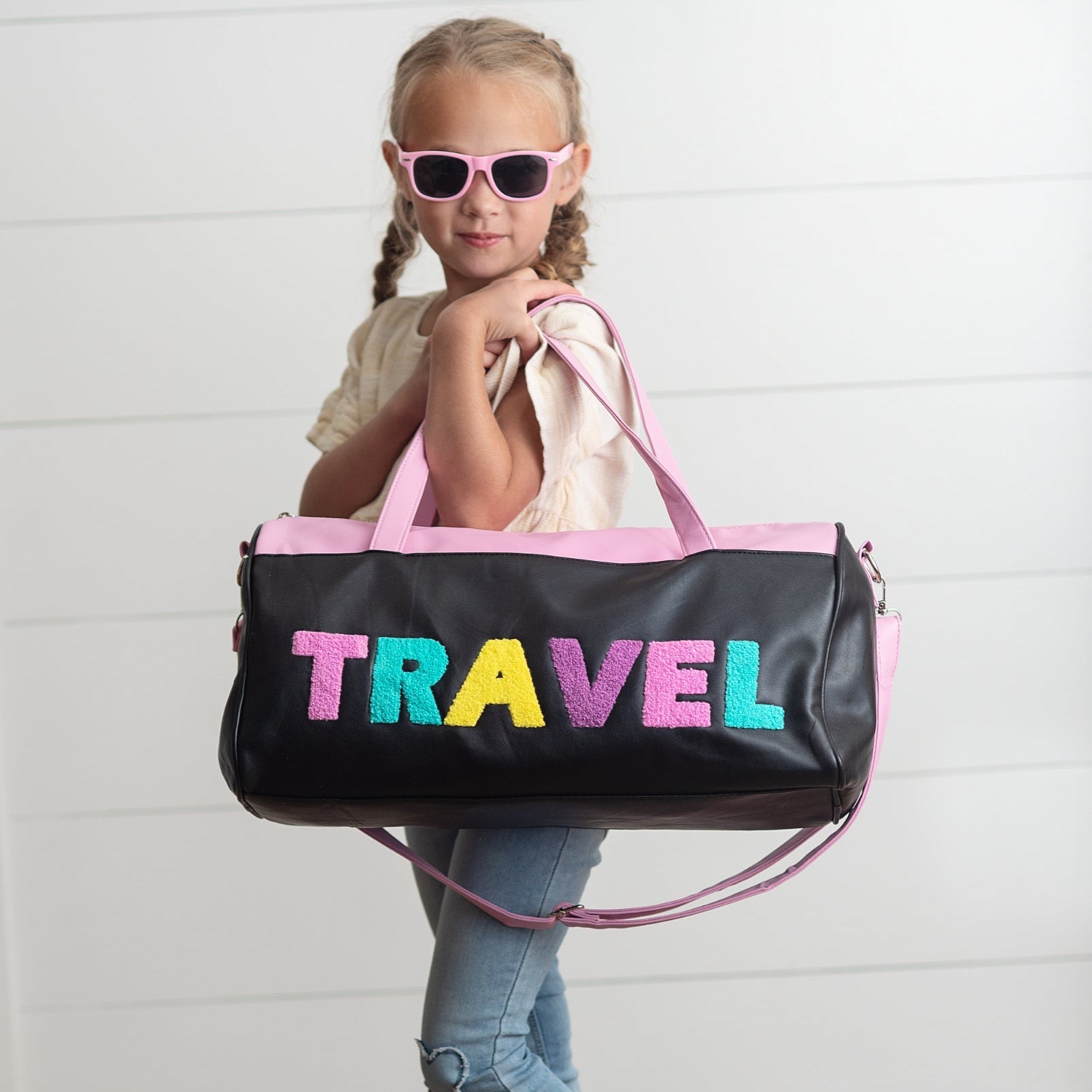 Child holding a black duffel bag with colorful 'TRAVEL' letters against a white background.