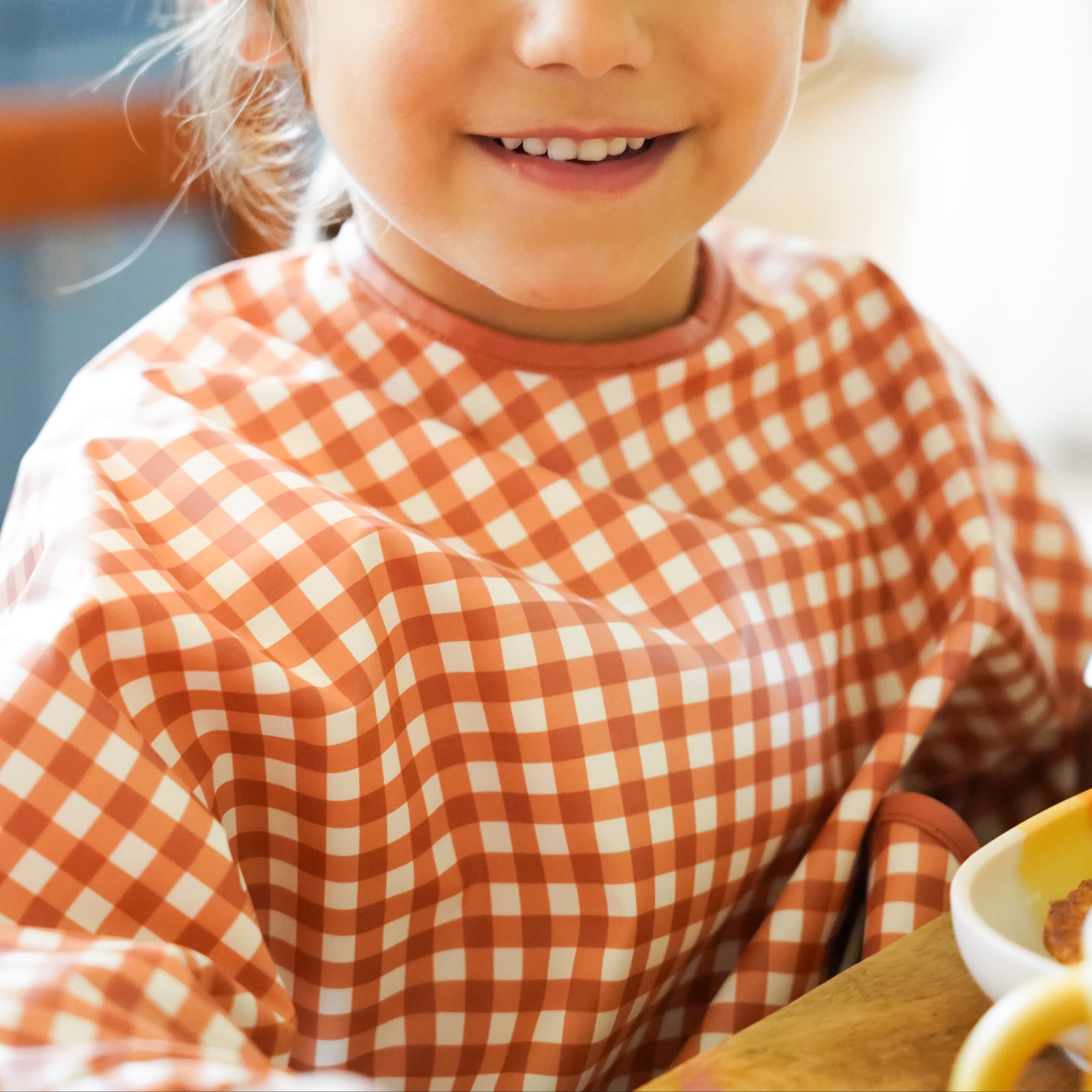 Young girl wearing a red and white checkered shirt, sitting at a table with a blurred background.