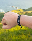 Black WWJD bracelet worn on a wrist outdoors overlooking green hills and water