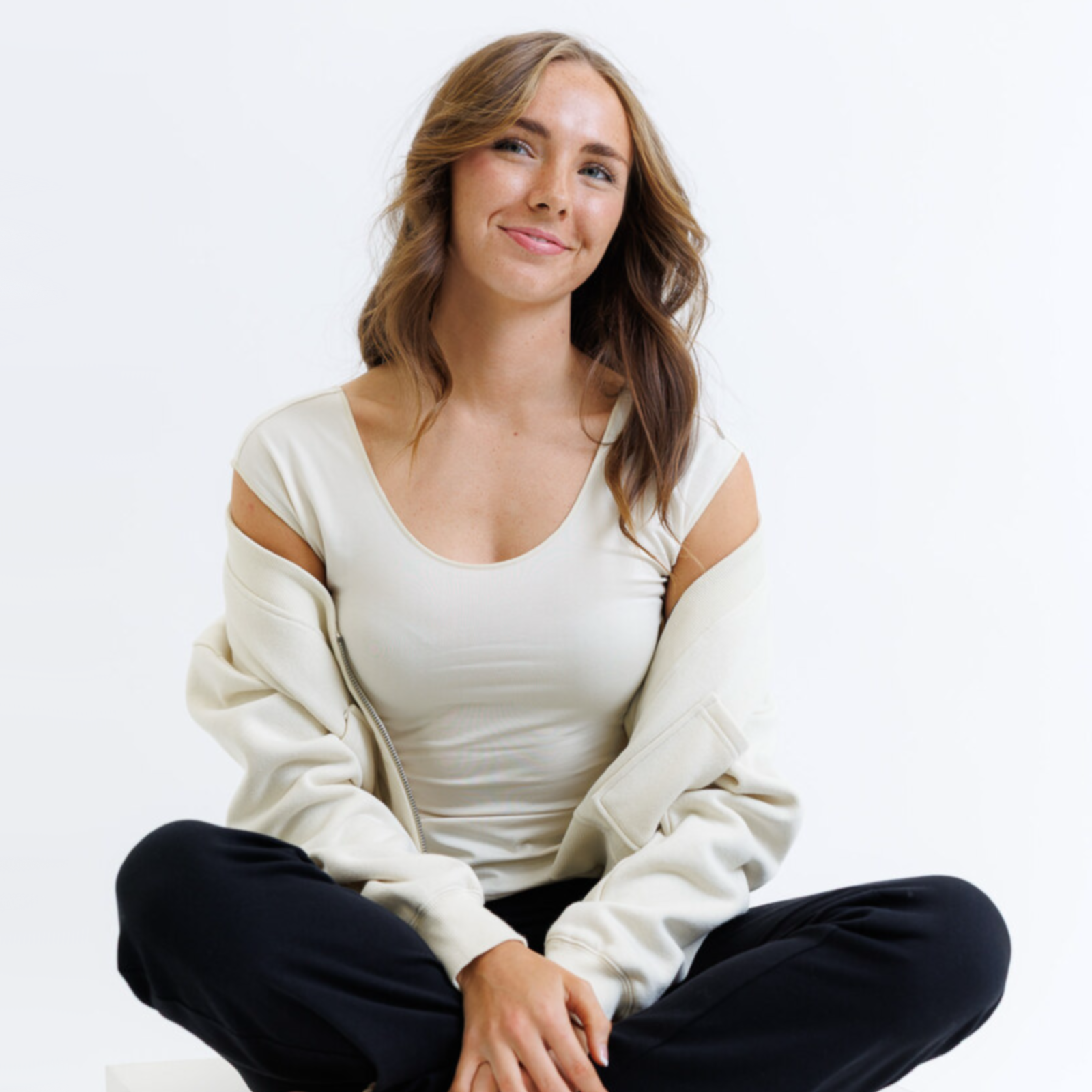 Woman sitting cross-legged on a white background wearing a white top and black pants.