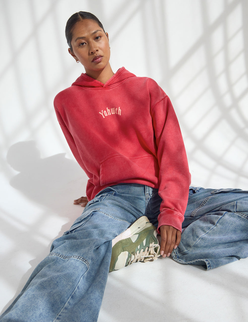 Woman seated in sunlight wearing Yahweh Ribbon Red Hoodie with a front pouch pocket