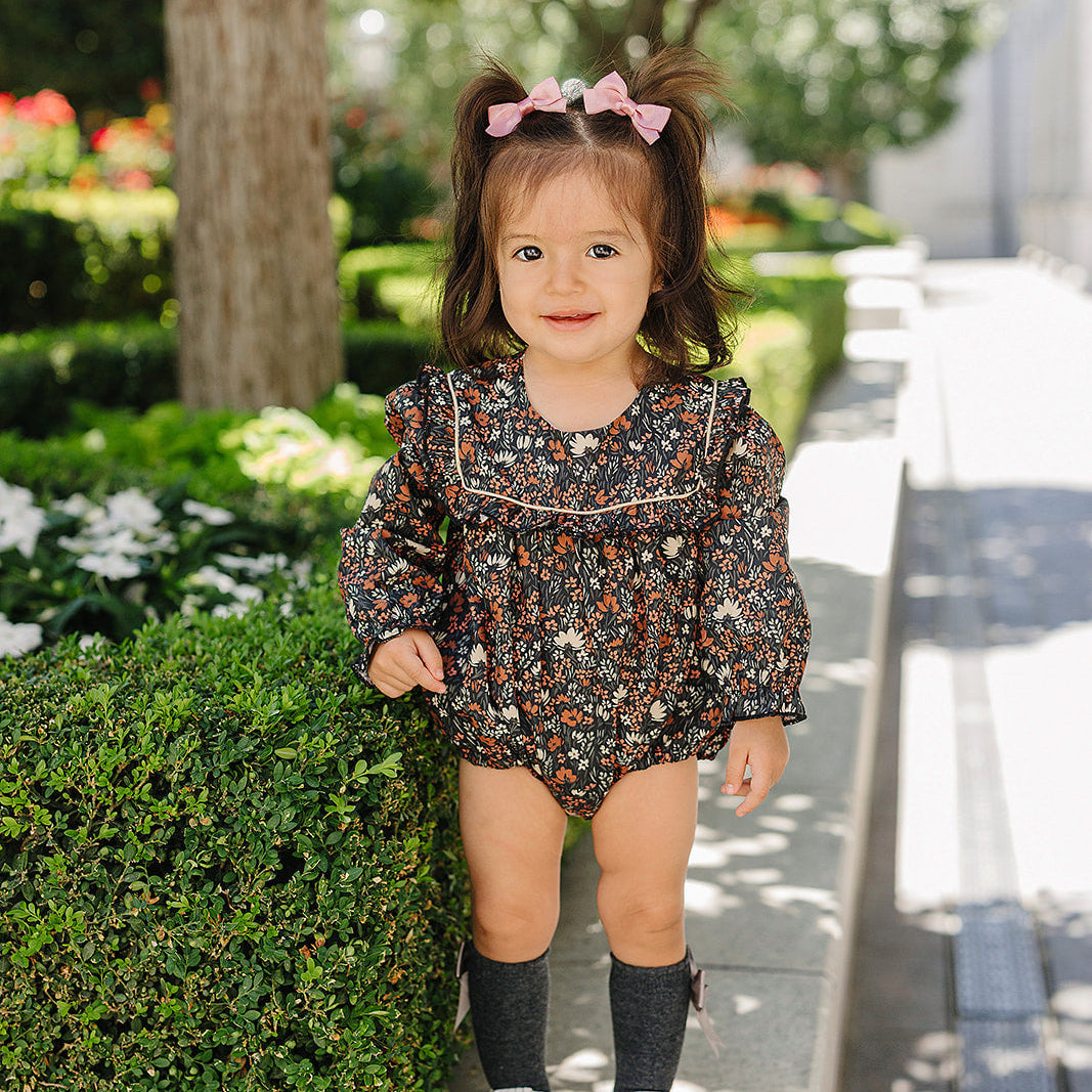 Young girl in a floral dress standing on a sidewalk with greenery in the background