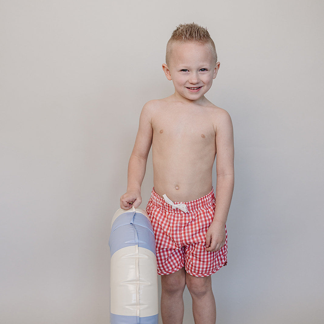 Young boy in red checkered swim trunks holding a colorful beach ball against a plain background