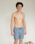 Man wearing blue checkered boxer briefs standing next to a woven basket on a plain background