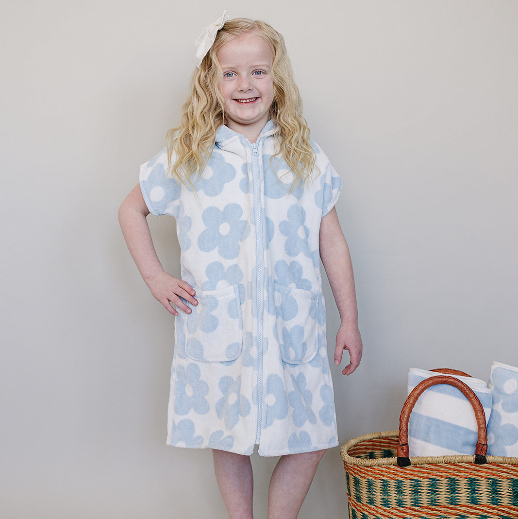 Young girl wearing a blue floral dress standing next to a woven basket with towels on a plain background
