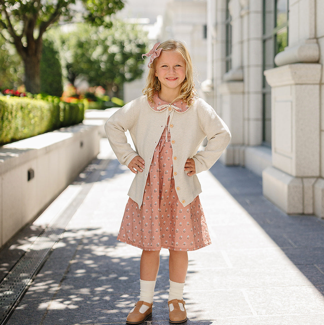 Young girl in a dress standing on a sunlit sidewalk with a building and trees in the background