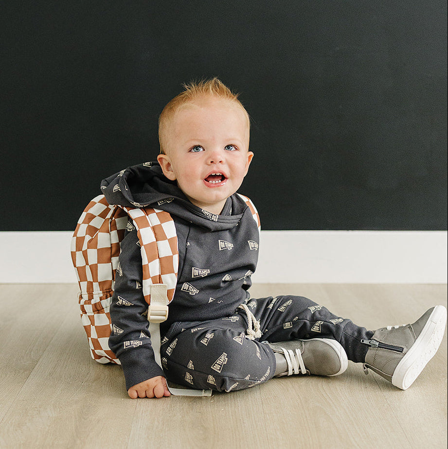 Baby sitting on a wooden floor wearing a checkered backpack against a black wall.
