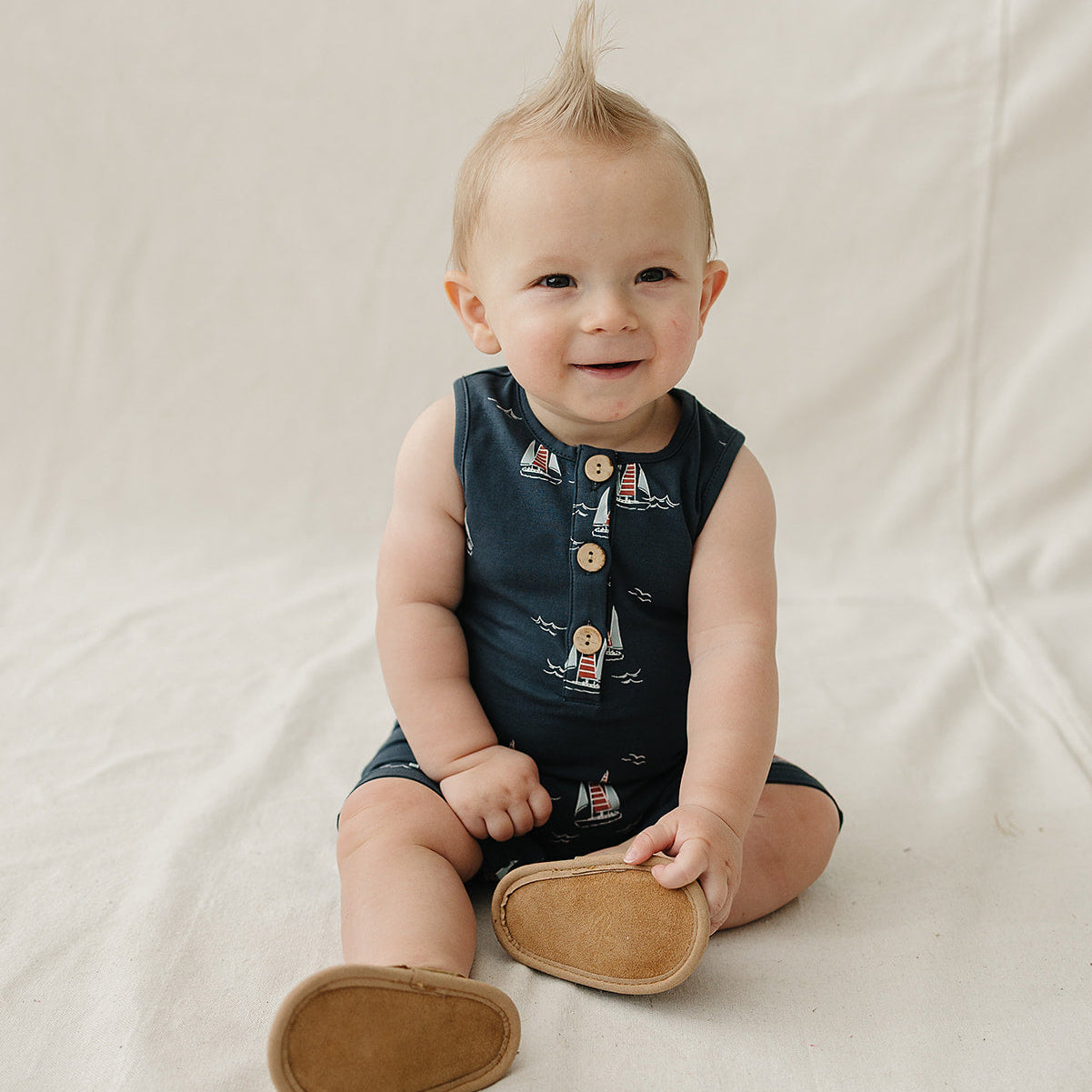 Baby sitting on a white blanket wearing a navy blue sleeveless dress with buttons and brown shoes.