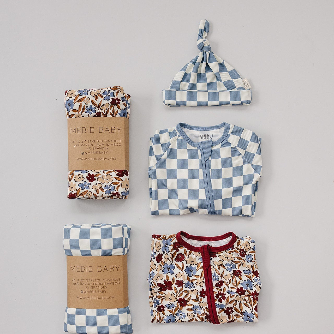 Collection of baby clothing and accessories including a floral blanket, checkered hat, and patterned outfit on a gray background.
