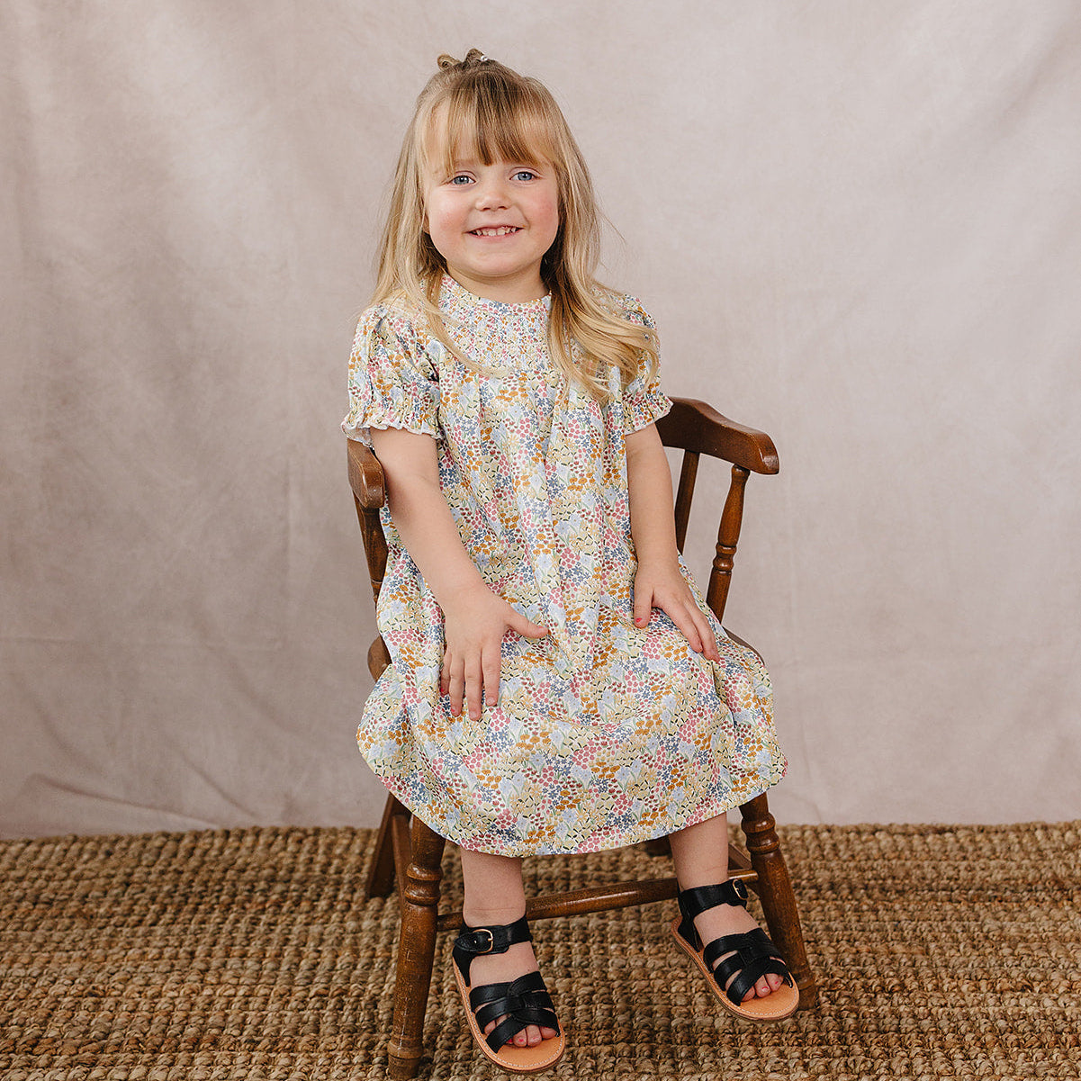 Young girl in a floral dress sitting on a wooden chair against a plain background