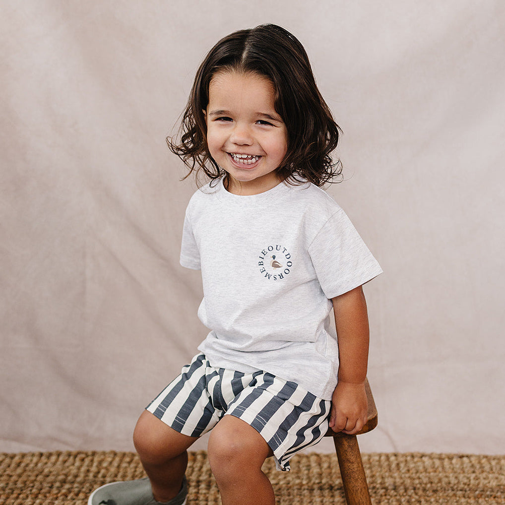 Child sitting on a wooden stool wearing a white t-shirt and striped shorts against a beige background