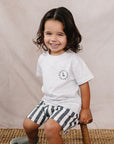 Child sitting on a wooden stool wearing a white t-shirt and striped shorts against a beige background