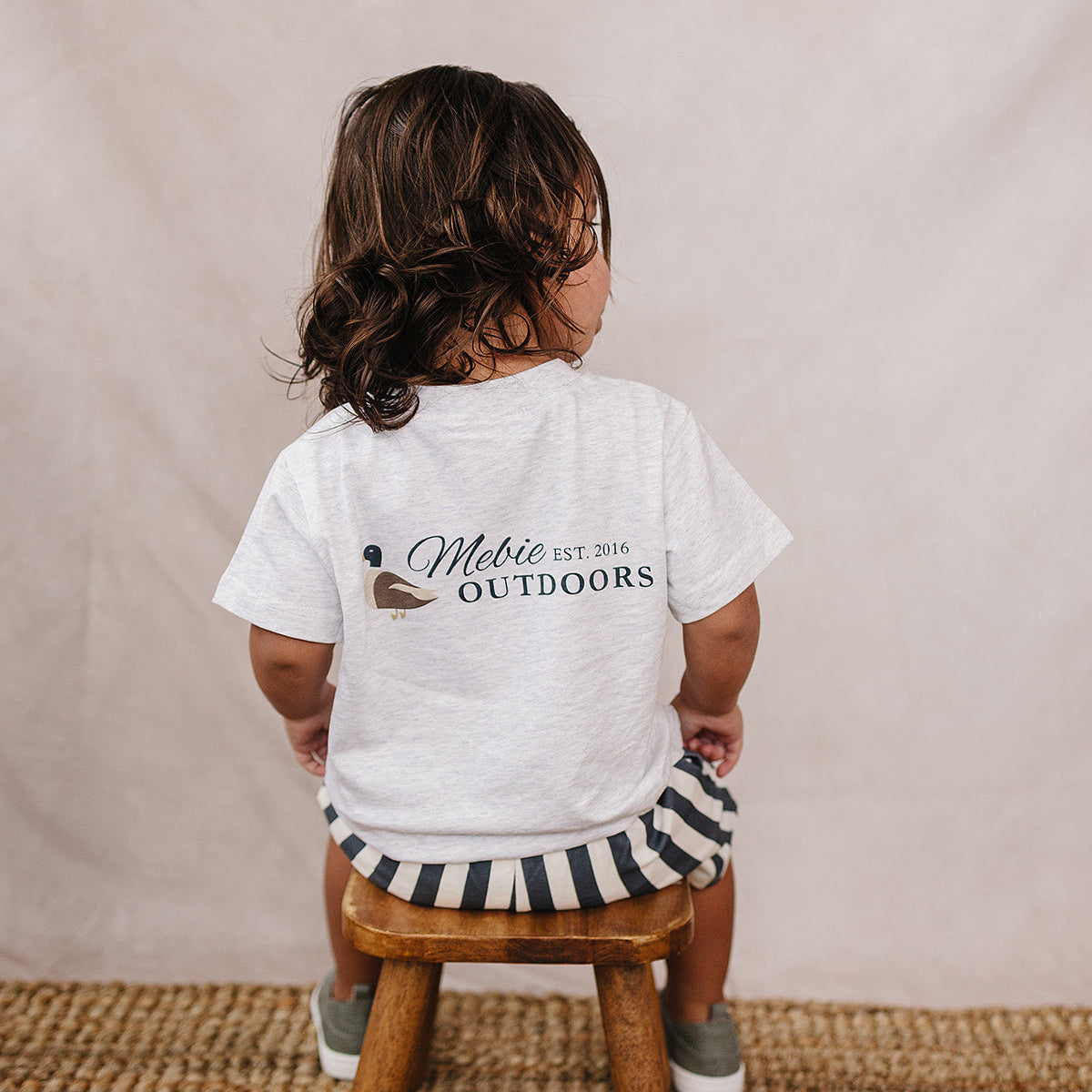 Child wearing a white t-shirt with 'Mebie Outdoors' logo, sitting on a wooden stool against a beige wall.