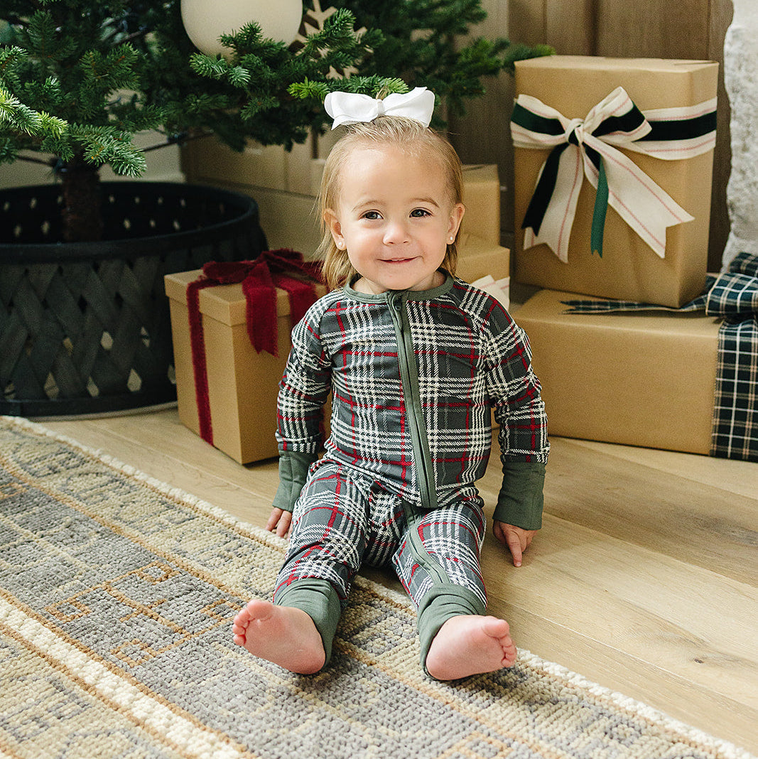 Child in plaid pajamas sitting on the floor with a Christmas tree and presents in the background