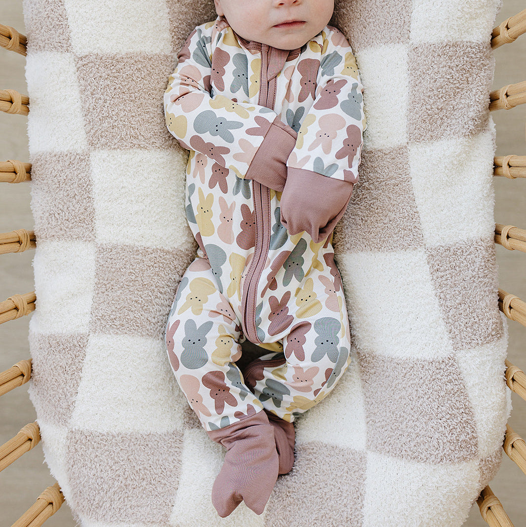 Baby in a floral onesie and bow headband lying on a checkered blanket in a wooden crib.