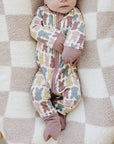 Baby in a floral onesie and bow headband lying on a checkered blanket in a wooden crib.