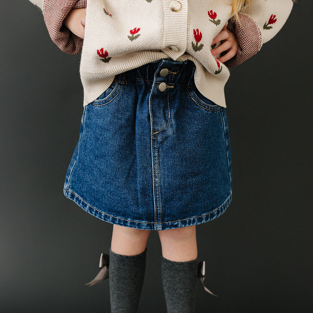 Person wearing a patterned cardigan, denim skirt, gray tights, and white shoes on a black background