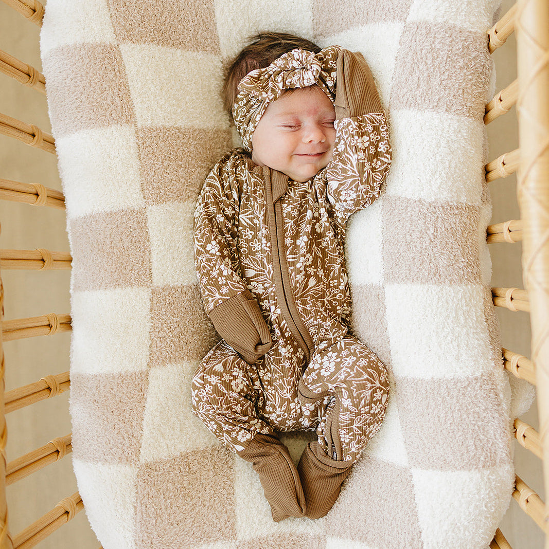 Newborn baby in a brown patterned outfit and headband lying on a checkered blanket in a wooden crib.