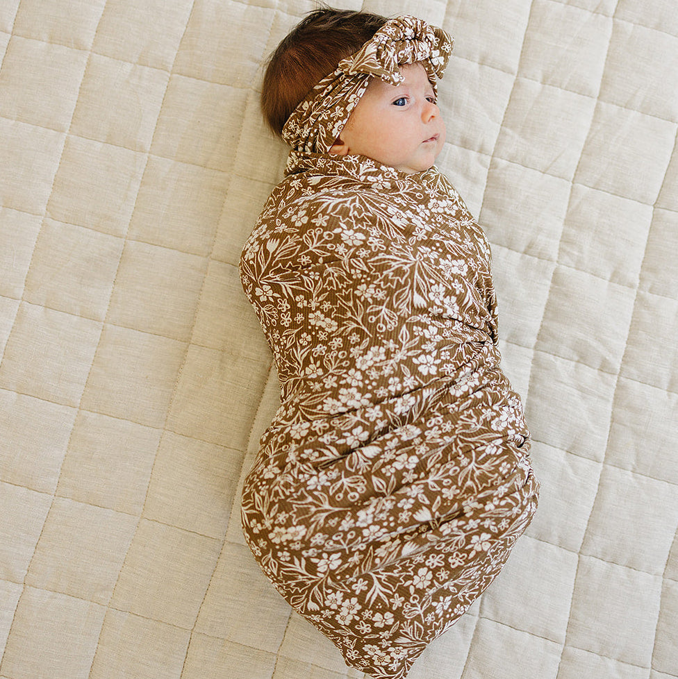 Baby wrapped in a floral swaddle and headband on a beige quilted surface