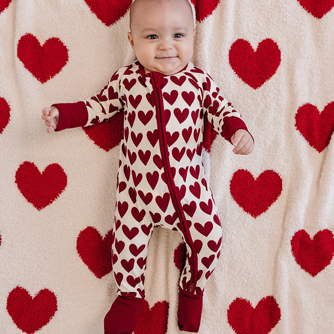 Baby in a heart-patterned onesie standing against a backdrop of red hearts on a white background