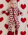 Baby in a heart-patterned onesie standing against a backdrop of red hearts on a white background