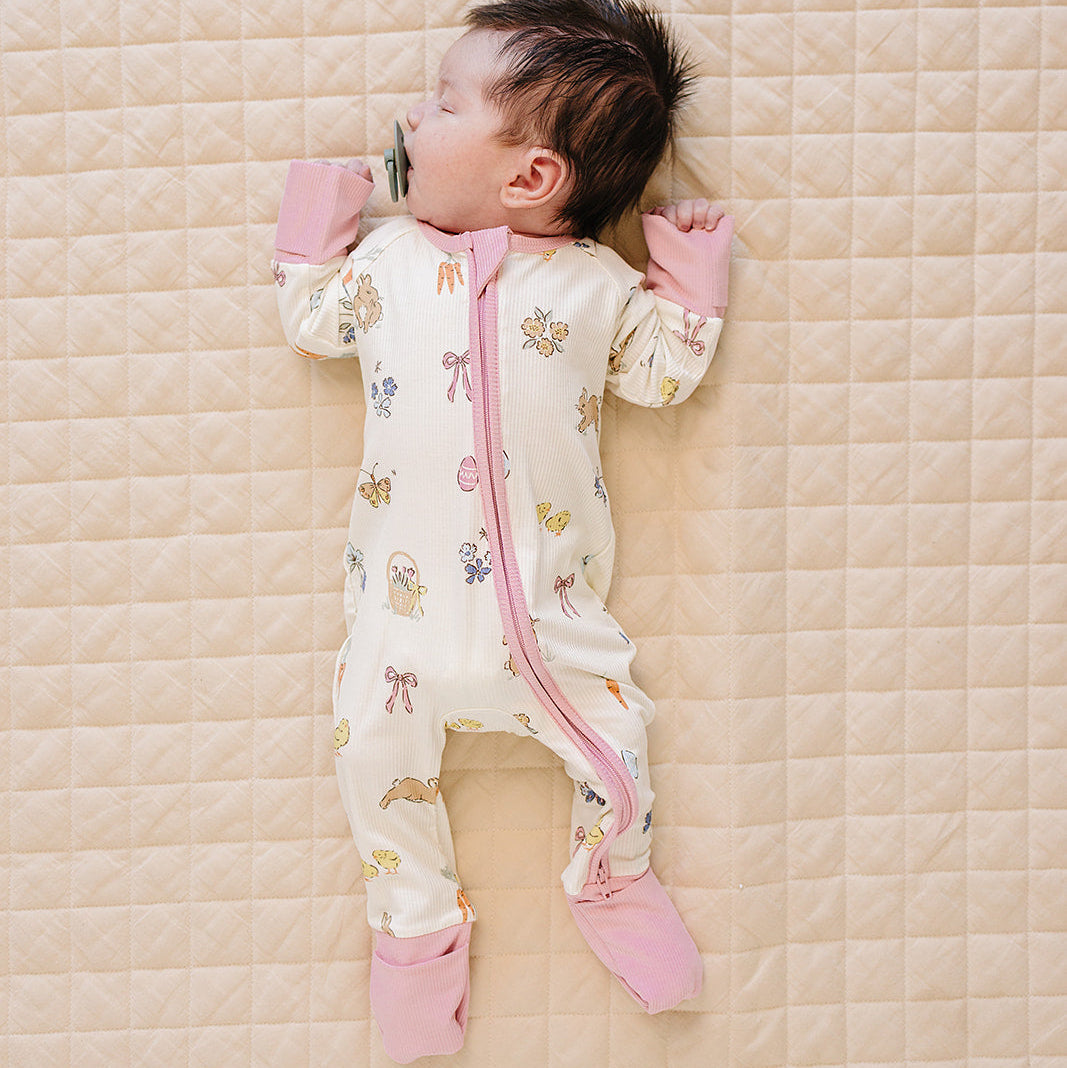 Baby lying on a quilted surface wearing a patterned onesie with pink accents.