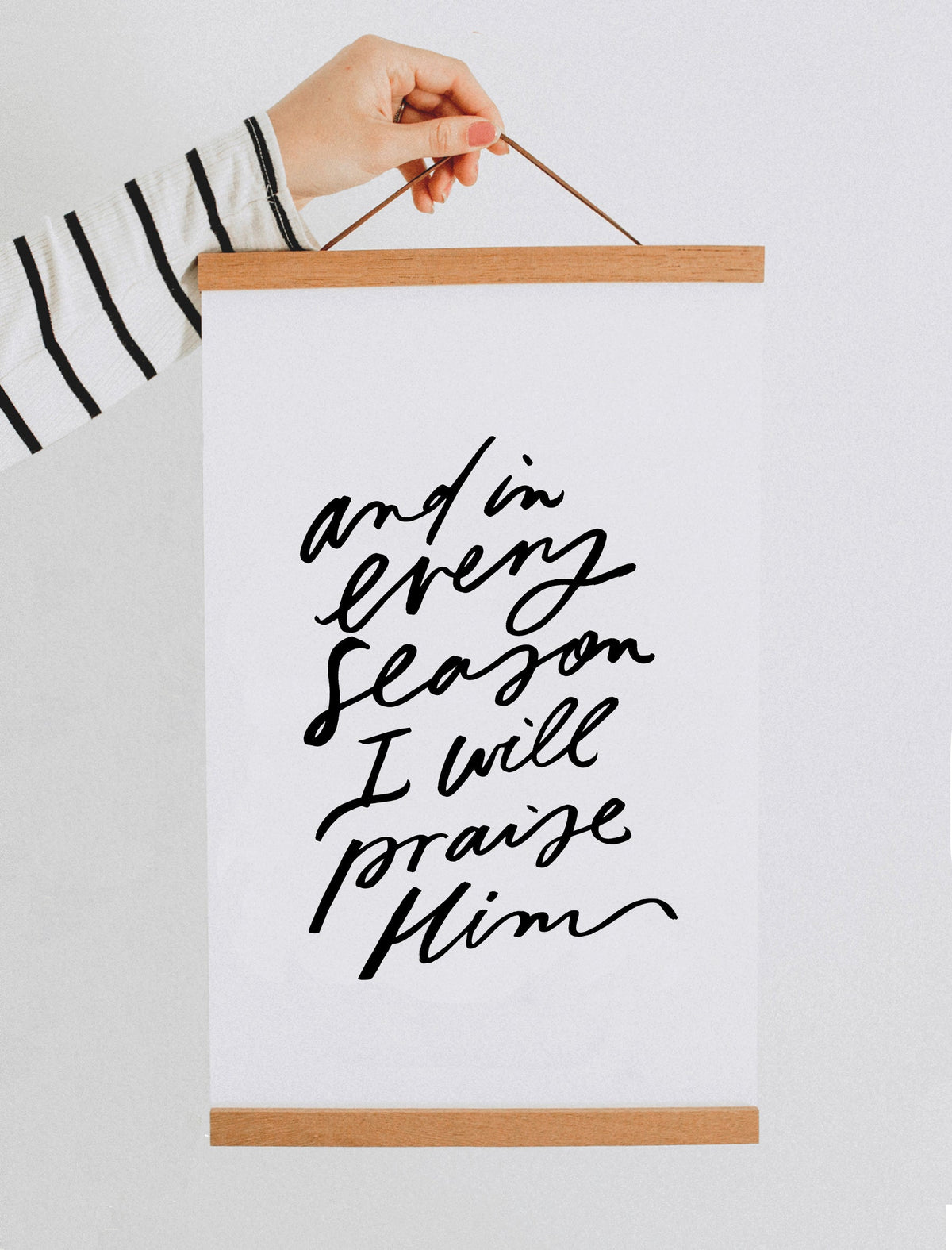And in every season I will praise Him art print with black script on white paper, displayed with a wood banner hanger held by a hand