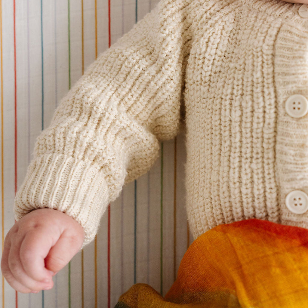 Child wearing a beige knitted sweater with colorful pants against a striped background