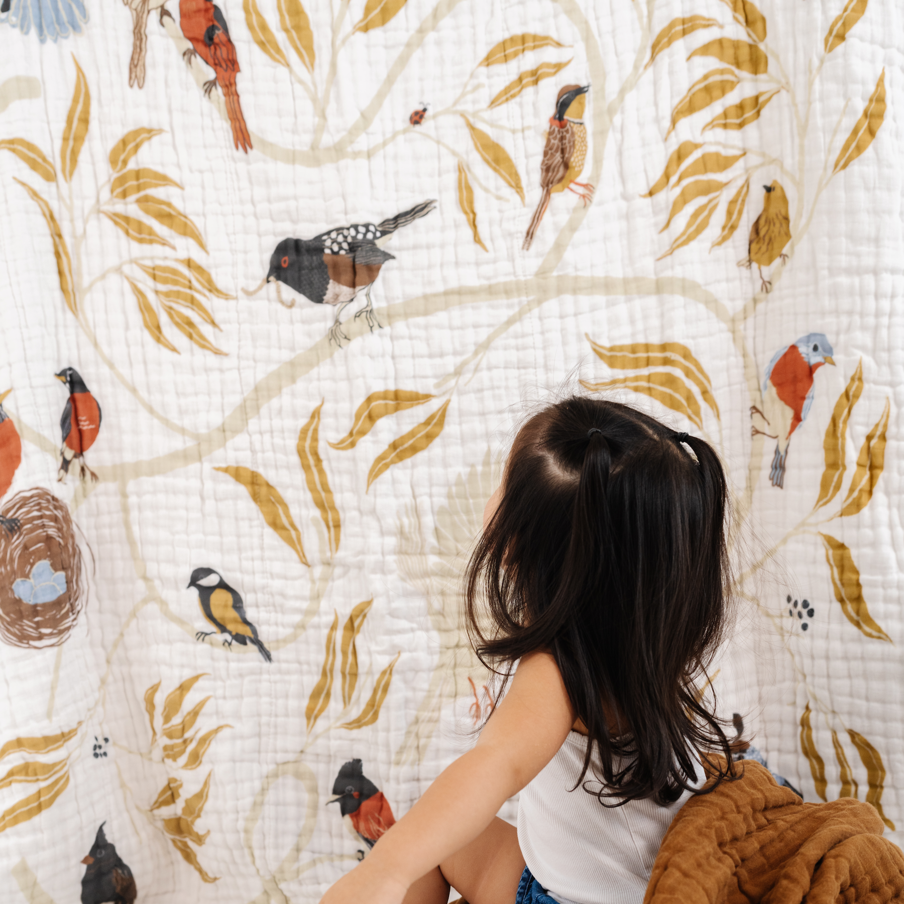 Child sitting on a white floor against a wall with a nature-themed quilt featuring birds and leaves.