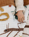 Person holding a quilt with dinosaur patterns, wearing a white sweater and brown pants.