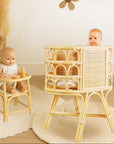 Two baby dolls in a rattan high chair and smaller stool with decorative flowers and a rabbit picture on the wall.