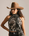 Woman wearing a brown cowboy hat, black floral halter top, and dark jeans against a beige background