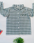 Green gingham boys’ shirt arranged flat with sleeves raised beside colored pencils and greenery.