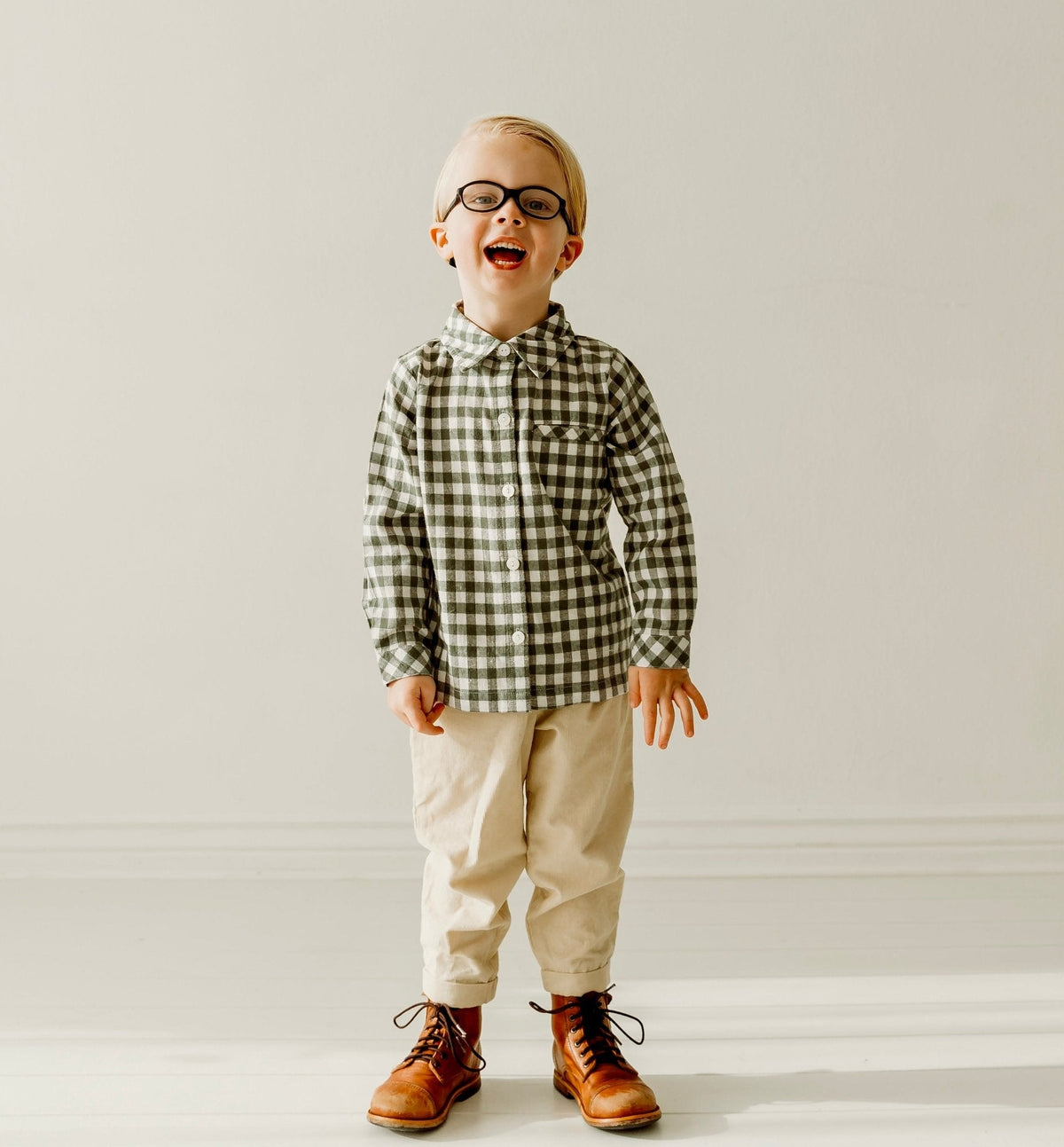 Boy wearing a green and white gingham long sleeve shirt with cream trousers and brown boots.