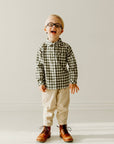 Boy wearing a green and white gingham long sleeve shirt with cream trousers and brown boots.